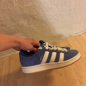 Blå Adidas Campus sneakers - Säljer ett par blå Adidas Campus sneakers med vita ränder och vit sula. Skorna har klassisk snörning och är tillverkade i mocka med gummisula. Perfekta för dig som vill ha ett par snygga skor i sommar! Blåa skosnören tillkommer! pris kan diskuteras 