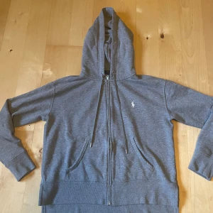 Grå hoodie från Polo Ralph Lauren - Säljer en klassisk grå hoodie från Polo Ralph Lauren med vit broderad logga på bröstet. Tröjan har dragkedja, huva med snörning och två fickor framtill. Perfekt för en avslappnad stil.