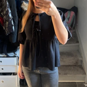 Svart blus med knyt och puffärm - Svart blus med knyt framtill och liten öppning vid halsen. Blusen har puffiga ärmar och en lös passform som ger en avslappnad men ändå söt look. Perfekt att styla med jeans eller kjol.