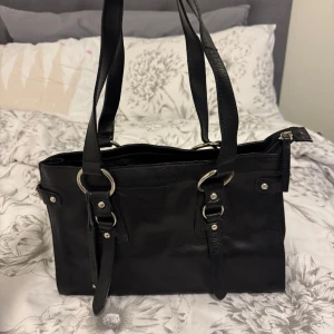 Svart handväska med spänndetaljer - Säljer en svart handväska med dubbla axelband och snygga spänndetaljer i silver. Väskan har dragkedja upptill och är rymlig inuti, perfekt för att bära med sig det viktigaste. Klassisk och stilren design som passar till många outfits.