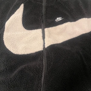 Svart fleecejacka från Nike med stor logga - Svart fleecejacka från Nike med dragkedja och stor vit Nike-logga över bröstet. Jackan har mjuk och fluffig yta, perfekt för kyligare dagar. Klassisk sportig stil med hög komfort.