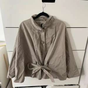 Beige poncho med knappar och knytband - Säljer en beige capejacka med krage, stora knappar framtill och knytband i midjan. Jackan har vida ärmar och en lös passform som ger en avslappnad och trendig look. Perfekt för lager-på-lager under höst och vår. I trenchcoat stuket.