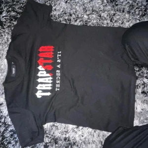 Svart Trapstar t-shirt med tryck - Svart t-shirt från Trapstar med stort tryck i vitt och rött på bröstet där det står 'TRAPSTAR' och 'IT'S A SECRET'. Klassisk passform och rund halsringning. Perfekt för en streetwear-look. (Nytt med kartongen på )