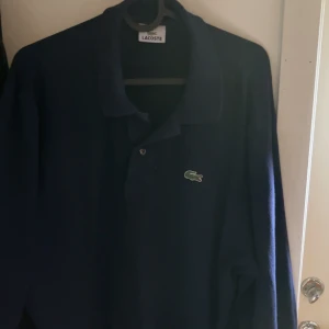 Mörkblå pikétröja från Lacoste - Säljer en klassisk mörkblå pikétröja från Lacoste med den ikoniska krokodilloggan på bröstet. Tröjan har krage och knäppning framtill, perfekt för en stilren look. Underbar nu till sommaren och varmare väder! Priset kan diskuteras vid snabb affär!