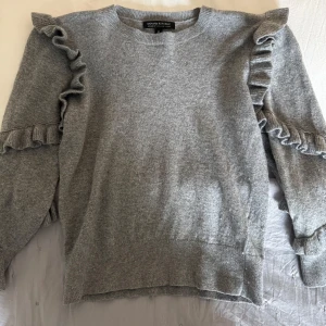 Grå volangtröja från Banana Republic - Superfin grå stickad tröja från Banana Republic med volangdetaljer längs axlar och ärmar. Mjuk och skön, perfekt för lager-på-lager. Rund halsringning och ribbade muddar. Tillverkad av italienskt garn.