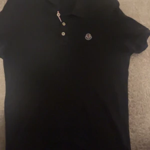 Svart pikétröja från Moncler - Säljer en klassisk svart pikétröja från Moncler med korta ärmar och knappslå framtill. Tröjan har Moncler-logga på bröstet och snygga detaljer vid knapparna. Perfekt för en stilren look.