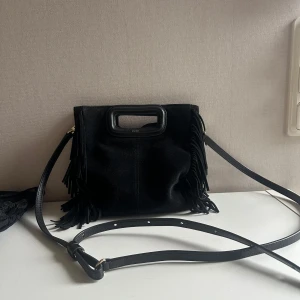 Svart axelväska med fransar från Maje - Säljer en svart axelväska från Maje med fransar på sidorna och ett stilrent handtag upptill. Väskan har justerbar axelrem och är tillverkad i mocka. Perfekt accessoar för att lyfta din outfit. Använd ett fåtal gånger så i nyskick!