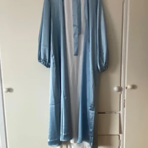 Abaya | Ljusblå långklänning med knytband - Säljer en elegant ljusblå långklänning med långa ärmar och knytband vid halsen. Klänningen har en lös passform och är tillverkad i ett glansigt, satinliknande material som ger en lyxig känsla. Den är helt ny. Passar längder 160-170cm!