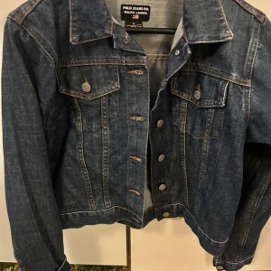 Blå jeansjacka från Polo Jeans Co. Ralph Lauren - Klassisk blå jeansjacka från Polo Jeans Co. Ralph Lauren med knappar framtill och två bröstfickor med lock. Jackan har en tidlös design med krage och långa ärmar. Perfekt att slänga på sig över en t-shirt eller hoodie.