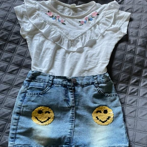 Jeanskjol med smiley-paljetter - Säljer en söt jeanskjol från UNS Wear i ljusblå denim med två stora guldiga smiley-paljetter framtill. Kjolen har klassisk femficksdesign, bälteshällor och knappstängning. Perfekt för dig som gillar unika detaljer!