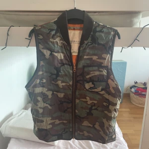 Camouflageväst med amerikansk flagga från Ralph Lauren - Säljer en unik väst från Ralph Lauren Denim & Supply med camouflage-mönster framtill och amerikansk flagga på ryggen. Västen har dragkedja, bröstficka och ribbad krage. Fodret är orange och materialet känns slitstarkt.