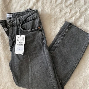 Grå jeansbyxor från Zara - Säljer ett par grå jeansbyxor från Zara med raka ben och slitningar vid knäna. Byxorna har klassisk femficksdesign, knapp och dragkedja i gylfen. Perfekta för en avslappnad och trendig look.