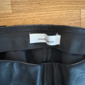 Svarta skinnbyxor från Samsøe Samsøe - Snygga svarta skinnbyxor från Samsøe Samsøe med minimalistisk design. Byxorna har en smal passform och är tillverkade i ett material som ser ut som skinn. Perfekta för en trendig och stilren look.