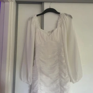 Vit kortklänning med långa ärmar och rynkade detaljer - Säljer en vit kortklänning med långa, luftiga ärmar och rynkade detaljer framtill. Klänningen har en fyrkantig ringning och dragkedja i ryggen. Använd 1 gång vid student! Säljer då den är för liten!❤️