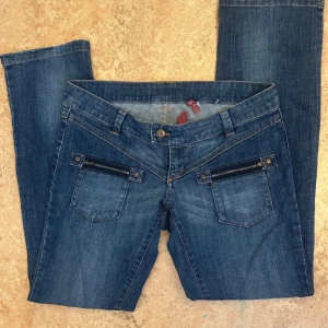Blå jeansbyxor med dragkedjedetaljer - Säljer ett par blå jeansbyxor med raka ben och två framfickor med dragkedjor. Byxorna har även bakfickor med knappdetaljer och klassisk jeanslook. Perfekta för en avslappnad stil.