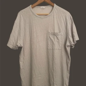 Filippa K Linne tshirt - En basic ljusgrå t-shirt från Filippa K med rund halsringning och en enkel bröstficka. Perfekt för en avslappnad stil och lätt att matcha med andra plagg.