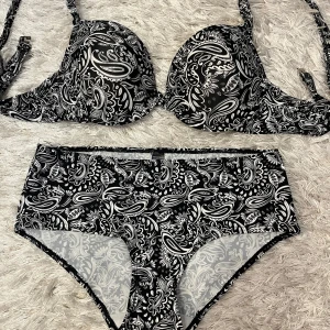 Svart och vit bikini från Ellos - Snygg bikini från Ellos med svart och vitt paisleymönster. Överdelen har vadderade kupor och knytband, medan underdelen är högmidjad. Perfekt för strand eller poolhäng!