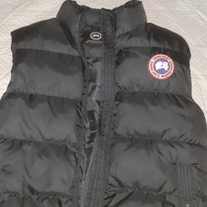 Svart dunväst från Canada Goose - Säljer en svart dunväst från Canada Goose med klassisk logotyp på bröstet. Västen har hög krage och stängs med dragkedja. Perfekt för lager-på-lager under kyliga dagar.