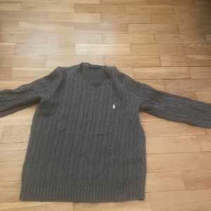 Grå stickad tröja från Ralph Lauren - Kabelstickad grå tröja från Ralph Lauren med v-ringning och diskret broderad logga på bröstet. Klassisk modell med ribbade muddar vid ärmslut och nederkant. Perfekt för lager-på-lager under kyliga dagar.