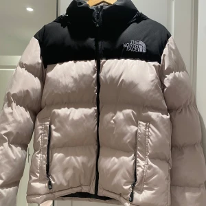 Beige och svart dunjacka från The North Face - Säljer en beige och svart dunjacka från The North Face med huva och dragkedja framtill. Jackan har två fickor med dragkedja och logga på bröstet. Aldrig använd.