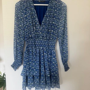 Blå mönstrad klänning från Zara - Supersnygg blå kortklänning från Zara med vitt och grönt mönster. Klänningen har v-ringning, långa ärmar med resår vid ärmslut och volangdetaljer i kjolen. Perfekt för dig som gillar färg och mönster!