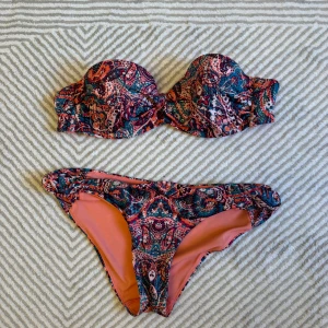 Mönstrad bandeau bikini - Säljer en färgglad bandeau bikini med paisleyinspirerat mönster i rött, blått och rosa. Överdelen är axelbandslös med lätt vaddering och underdelen har en klassisk bikini-trosa modell. Perfekt för stranden eller poolen!