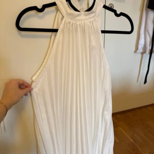 Vit plisserad långklänning med knyt i nacken - Säljer en elegant vit långklänning med plisserat tyg och hög hals. Klänningen har knytband i nacken och är ärmlös, vilket ger en stilren och luftig. Klänningen har dubbeltyg och därav inte genomskinlig. Klänningen är köpt i Spanien. Oanvänd klänning.