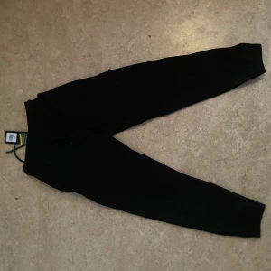 Svarta mjukisbyxor från Under Armour - Svarta mjukisbyxor från Under Armour med logga på låret och text längs benet. Byxorna har resår i midjan med snörning och muddar vid bensluten. Perfekta för träning eller chill.