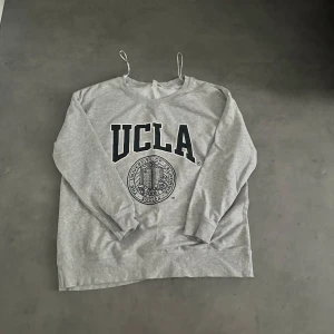 Grå sweatshirt med UCLA-tryck från H&M - Säljer en grå sweatshirt från H&M med stort svart UCLA-tryck och universitetslogga framtill. Tröjan har rund halsringning och långa ärmar. Perfekt för en avslappnad och sportig stil.
