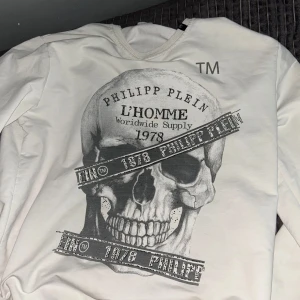 Vit långärmad tröja med dödskalle från Philipp Plein - Säljer en vit långärmad tröja från Philipp Plein med stort tryck av en dödskalle och texten 'L'HOMME Worldwide Supply 1978' på framsidan. Tröjan har ribbade muddar och en cool streetstil.