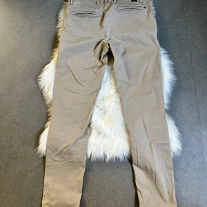 Beige chinos från Replay - Snygga beige chinos från Replay med raka ben och klassiska fickor bak med dragkedja. Byxorna har bälteshällor och är perfekta för en clean och stilren look. Sköna och lätta att matcha med det mesta.