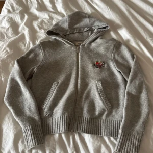 Zip hoodie från Zadig & Voltaire - Säljer en grå hoodie från Zadig & Voltaire med dragkedja och huva. Tröjan har ribbade muddar, två fickor framtill och ett litet broderat märke på bröstet. Perfekt för en avslappnad stil.
