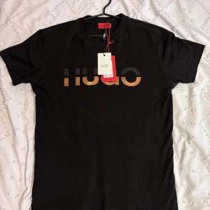  Hugo Boss T-shirt - Säljer en svart t-shirt från Hugo med stor metallicfärgad logga framtill. Klassisk rund halsringning och korta ärmar. Perfekt för dig som gillar stilrena plagg med en twist.