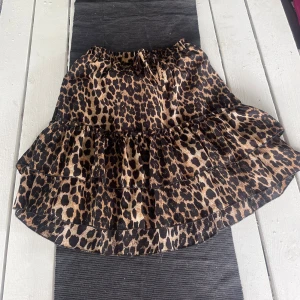 Leopardmönstrad volangkjol med knytband - Säljer en kort kjol med leopardmönster och volanger. Kjolen har resår i midjan och ett knytband framtill. Perfekt för dig som vill sticka ut med ett djurmönster och gillar volangdetaljer.