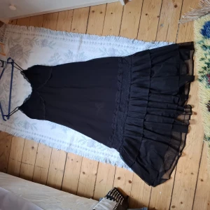 Svart långklänning med volang - Svart långklänning med smala axelband och volangdetaljer nertill. Klänningen har en feminin siluett och är tillverkad i ett lätt, transparent material som ger ett luftigt intryck. Perfekt för dig som gillar en stilren och elegant look.