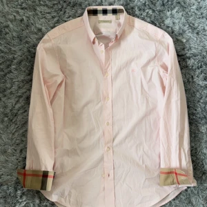 Ljusrosa skjorta från Burberry - Säljer en klassisk ljusrosa skjorta från Burberry med långa ärmar och ikoniska rutiga detaljer vid manschetterna och insidan av kragen. Skjortan har knappar framtill och är tillverkad i bomull med en liten andel elastan för extra komfort. Nypris idag ligger på 4800kr.