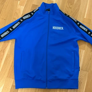 Blå track jacket från Hoodrich - Säljer en blå track jacket från Hoodrich med vit logga på bröstet och svarta band med vit text längs ärmarna. Jackan har dragkedja framtill, ståkrage och två fickor. Perfekt för en sportig stil. 