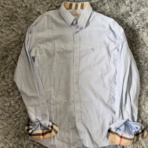 Ljusblå skjorta från Burberry med rutiga detaljer - Säljer en ljusblå skjorta från Burberry med klassiska rutiga detaljer på insidan av kragen och ärmsluten. Skjortan har lång ärm, bröstficka och knäppning framtill. Tillverkad i 100% bomull. Perfekt för dig som gillar stilrena plagg med en twist.