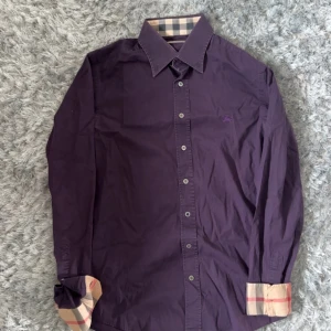Lila skjorta från Burberry med rutiga detaljer - Säljer en snygg lila skjorta från Burberry med klassiska rutiga detaljer på krage och ärmslut. Skjortan har knappar framtill och är långärmad. Perfekt för dig som gillar stilrena plagg med en twist.