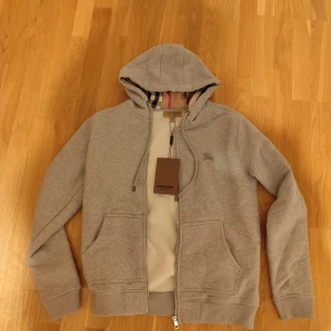 Grå hoodie från Burberry med dragkedja - Säljer en grå hoodie från Burberry med klassiskt rutigt foder på insidan. Tröjan har dragkedja, huva med snörning och två fickor framtill. Perfekt för dig som gillar stilrena och exklusiva plagg.