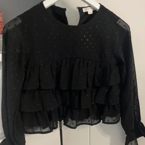 Svart volangblus från Gina Tricot - Säljer en svart blus från Gina Tricot med flera volanglager och små glittriga prickar. Blusen har lång ärm och rund halsringning, perfekt för att lyfta en outfit med lite extra detaljer. Säljer denna då den är för liten🫶🏼