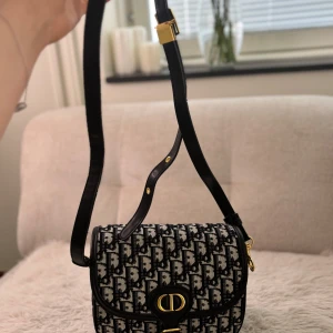 Dior 30 Montaigne axelväska i svart och vit canvas - Säljer en Dior 30 Montaigne axelväska i svart och vit canvas med ikoniskt Dior-mönster. Väskan har guldfärgade metalldetaljer, justerbar axelrem i svart läder och ett klassiskt CD-spänne framtill. Perfekt för dig som vill ha en lyxig accessoar med tidlös design.