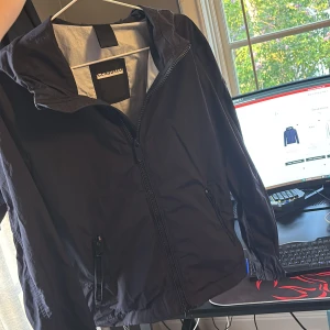 Svart vindjacka med huva från Censured - Säljer en svart vindjacka från Censured med huva och dragkedja framtill. Jackan har två fickor med dragkedja och en enkel, stilren design. Perfekt för blåsiga dagar och enkel att matcha med olika outfits.