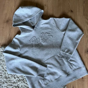 Grå hoodie från Polo Ralph Lauren - Säljer en stilren grå hoodie från Polo Ralph Lauren med broderad logga framtill. Tröjan har huva, lång ärm och en klassisk känguruficka. Perfekt för en avslappnad och trendig look.