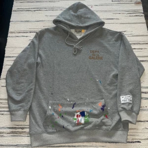 Grå hoodie från Gallery Dept. med färgstänk - Säljer en grå hoodie från Gallery Dept. med trycket 'DEPT. de la GALERIE' på bröstet och färgstänk på fickan och baksidan. Huvan har dragsko och ärmen har en patch med text. Perfekt för dig som gillar streetwear med unik touch.