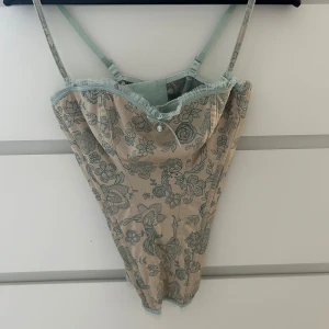 Blommig korsett från Marks & Spencer - Supersnygg korsett i ljusbeige med ljusblått blommönster och spetsdetaljer från Marks & Spencer. Justerbara axelband och liten rosett framtill. Är i väldigt bra skick och har endast blivit provad. 