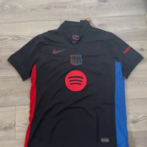 FC Barcelona svart bortatröja med Lamine Yamal 19 - Säljer en svart FC Barcelona bortatröja från Nike med Lamine Yamal och nummer 19 på ryggen. Tröjan har röda och blå detaljer på sidorna, klubbmärke och Nike-logga på bröstet samt Spotify-logga framtill.