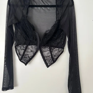 Svart mesh bolerotopp med spetsdetaljer - Säljer en svart bolerotopp i mesh med långa ärmar och vacker spets framtill. Toppen har öppen front och transparent material som ger en cool och trendig look. Perfekt att styla över ett linne eller klänning.