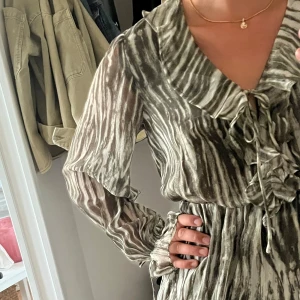 Grönmönstrad omlottklänning med volang - Säljer en kort klänning med grön- och vitmönstrat tyg, omlott och volangdetaljer framtill. Klänningen har långa ärmar och knytning vid halsen.