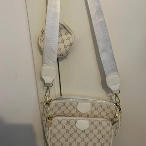 Vit axelväska med mönster och bred rem - Säljer en snygg vit axelväska med beige mönster och bred vit axelrem. Väskan har guldfärgade detaljer, dragkedja och en liten rund miniväska som sitter fast på remmen. Perfekt för att bära det viktigaste med stil.